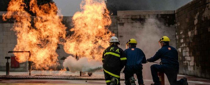 Three firefighters in full protective gear using a portable fire extinguisher to suppress controlled flames during a live fire simulation training exercise.
